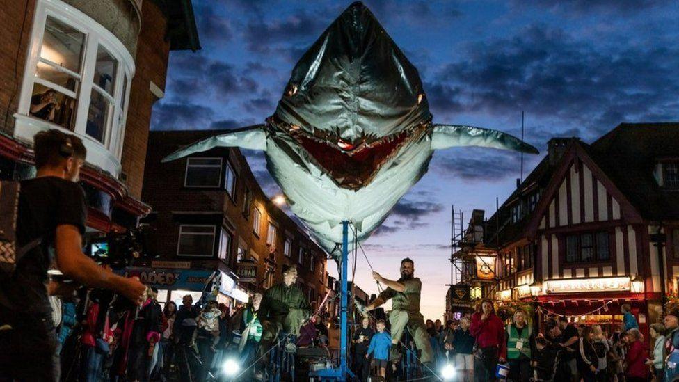 A giant metal sculpture of a shark moving through Weymouth's streets at night with crowds watching on