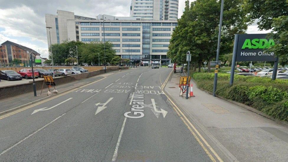 A city centre road. To the right is a sign marked 'Asda Home Office' and to the left is a car park. In the middle is a three-lane road with markings indicating it is one-way. A block of flats stands in the distance.