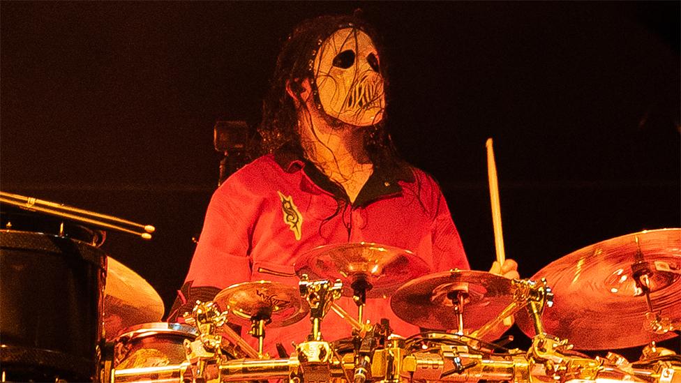 Jay Weinberg of Slipknot performs on stage in Finland. He is wearing a skull mask covering his face, with an orange tshirt which has a black collar. He has a wooden drum stick in his left hand, with the the drums he is playing are being shiny. He is looking out past the camera with the background black.