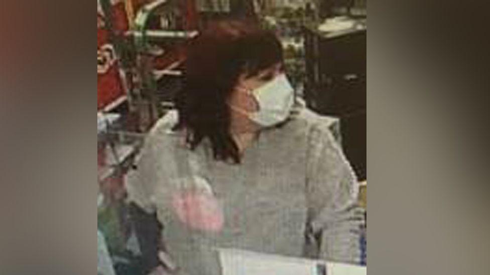 A woman with shoulder-length dark red hair wearing a Covid-style white face mask standing at a till. She is looking to her right and wearing a grey sweater