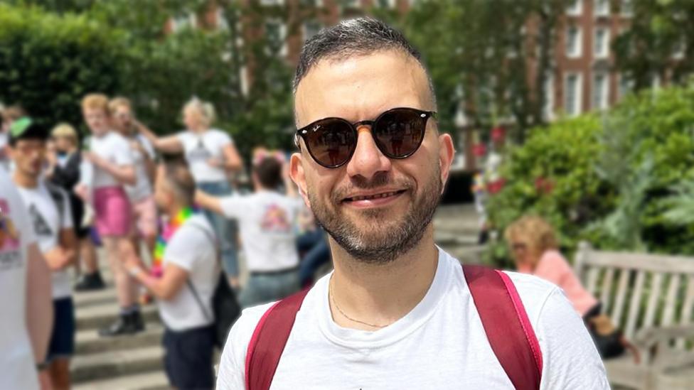 Giancarlo wearing sunglasses and smiling at the camera. He is wearing a white t-shirt and red backpack. The background is blurred, with trees and green hedges visible, along with multiple people.