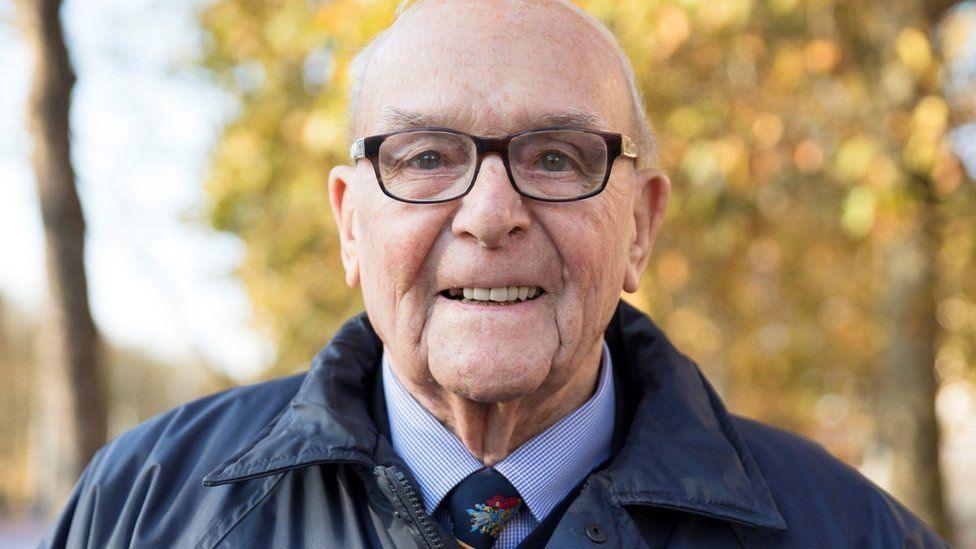 Roy Hayward smiling and looking straight at the camera. He is wearing glasses, a dark blue coat with shirt and tie.