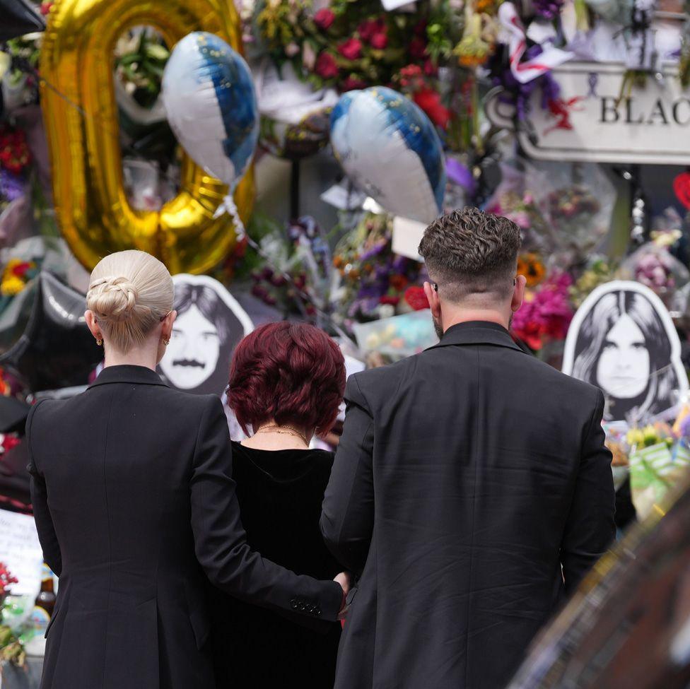 View of Kelly, Sharon and Jack from behind looking at a wall of colourful balloon and floral tributes, Kelly with her arm around Sharon's waist