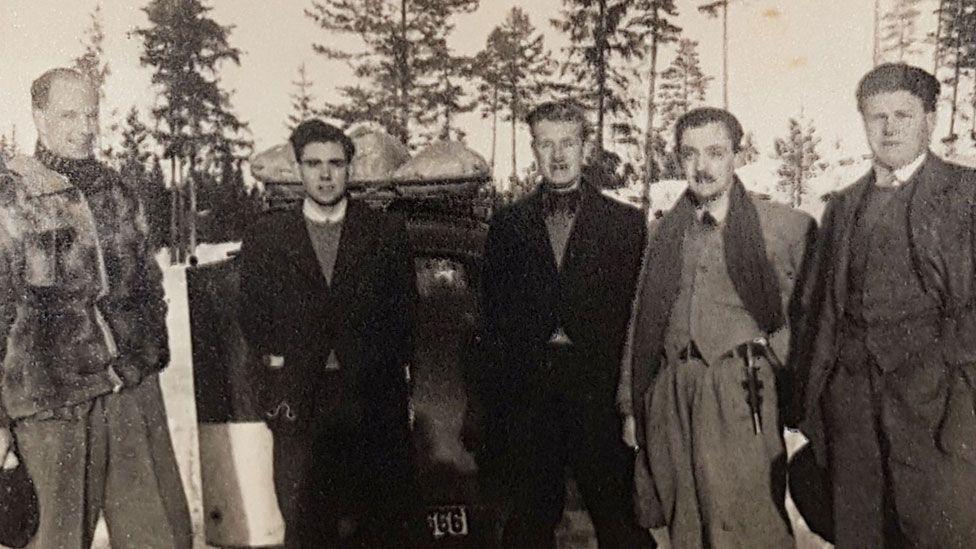 A black and white wartime photo of five men from left to right an unknown resistance worker, Michael Hicks, Robert Dalton, James Kenneth Bolton and Robert Chapin. They are lined up in front of a car and behind them is deep snow and pine trees outlined against the sky.