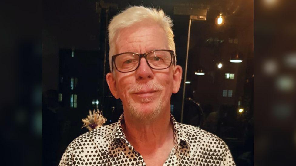 David Lloyd wearing a black and white patterned shirt and black framed glasses. He has short white hair and a trimmed white goatee. He is sitting in a restaurant at night with glass doors behind him, smiling at the camera.