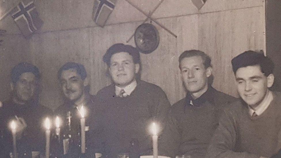 A close up of a black and white wartime photo showing Einar Thronsen, James Bolton, Robert Chapin, Robert Dalton and Michael Hicks sitting at a table with candles lit in front of them. Behind them is a wall and two Norwegian flags can be seen pinned to the walls.