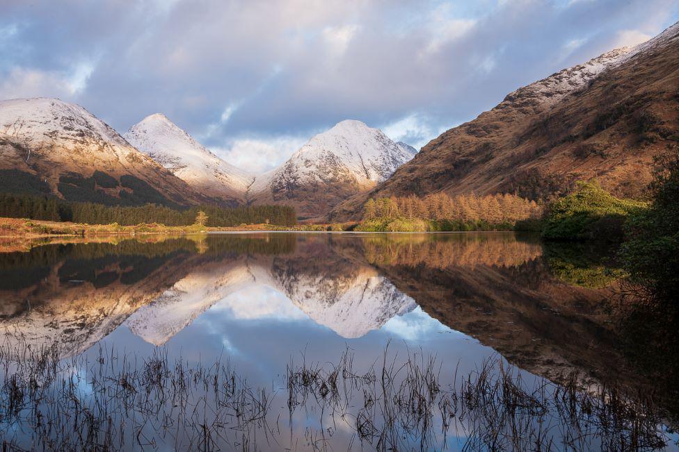 Snow-capped hills, and green/brown trees, reflected in water.