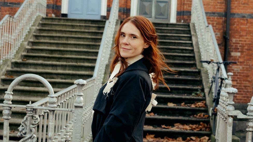 A woman with long red hair stands in front of a broad outdoor staircase with ornate white railings, set against a red-brick building. She is turned slightly toward the camera and wears a dark coat with light-coloured ribbon details. Fallen leaves are scattered on the steps.