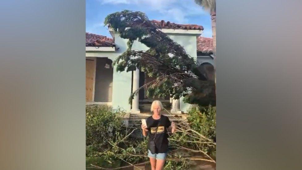 Video still of a woman with blonde hair, wearing a dark t-shirt and denim shorts, holding a mobile phone. She is standing in front of a building in pale green stucco, with a pan tiled roof and pillars in front of entrance. There is evidence of hurricane damage, with branches lying on the ground, and a tree is leaning at an angle above her head.