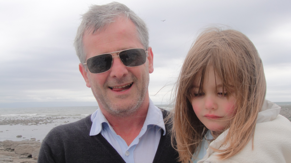 Manon Chiswell with her father Huw. Huw is looking at the camera and has his mouth open. He is wearing a blue shirt with a dark-grey cardigan over the top. He has black shades on and grey hair. He is holding his daughter, who is in the right of the photo, she has light-brown hair, and is wearing a white top. The sea and beach are behind them.