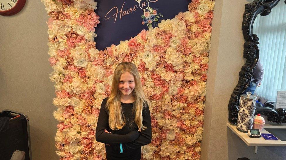 Lily-Mae before her charity haircut standing in front of a flower wall