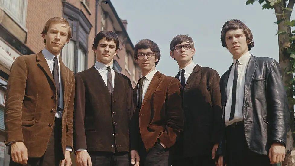 The original The Zombies photographed in the 1960s. They are wearing brown and black suade or leather jackets, dark ties and white shirts and have typical 1960s hairstyles. They are standing on a high street looking down at the camera which is tilted towards the sky, with a small tree on the right.