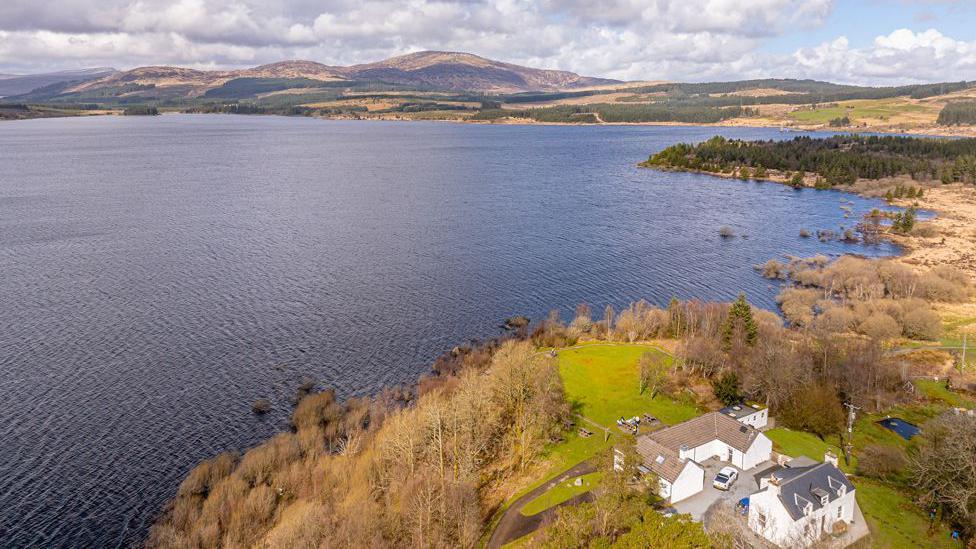 A gathering of white buildings next to a large loch with hills in the background