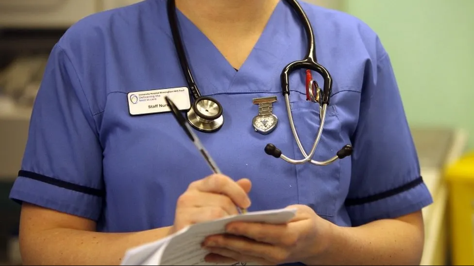 A hospital nurse in blue scrubs holding a pen and paper on a ward