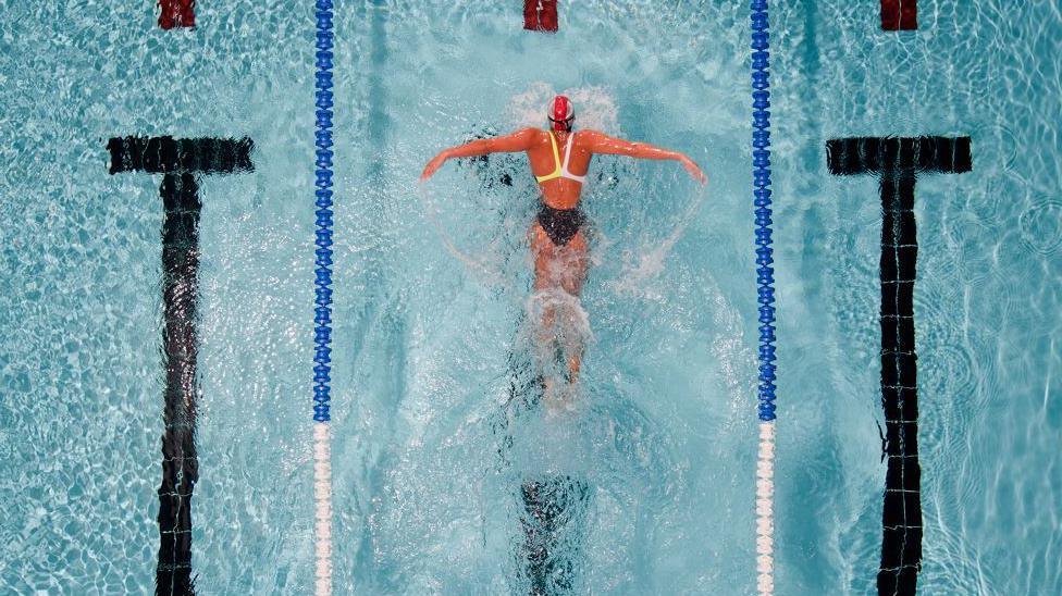 A lone woman in a swimsuit and swim cap doing butterfly in the middle lane of a swimming pool with water splashing up from the movement of her arms and legs