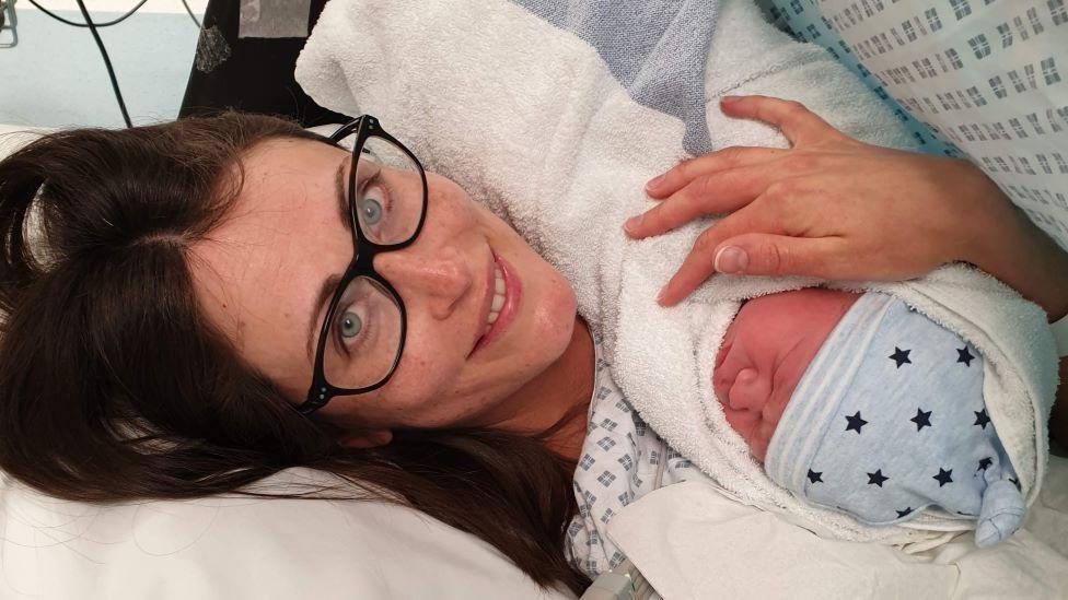 A woman with brown hair and glasses lies in a hospital bed, holding a newborn baby