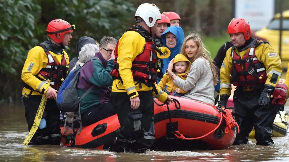 People are rescued in a boat from a flooded village by trained workers. In the boat you can see a family including a very young child.