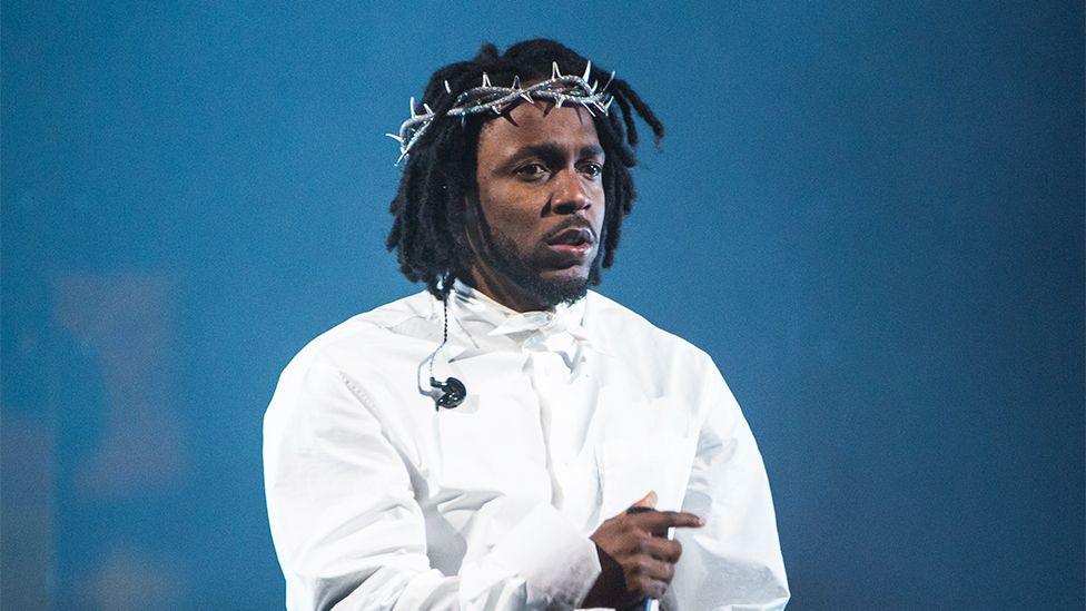 Kendrick Lamar, wearing a white shirt with a tiara-like crown around his forehead, performing on stage.