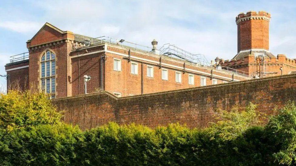 Red brick wall surrounds large building with long glass window and end, multiple windows at height and a turret