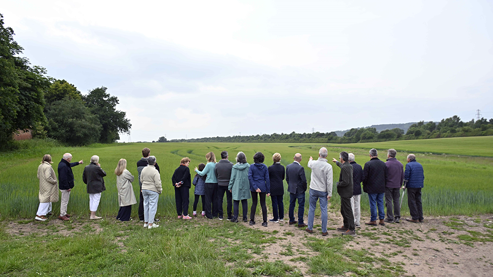 A group of 20 people looking away from camera at a green field, with hedgerows in distance. A few tall trees border the field on the left of the photo