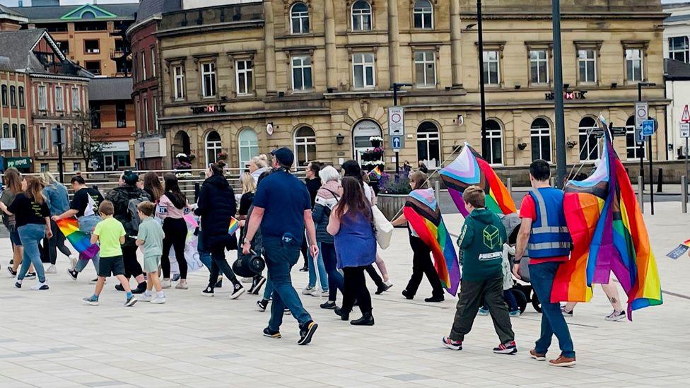 Parents and children dressed in casual clothing walk in a parade carrying rainbow flags through Rochdale town centre