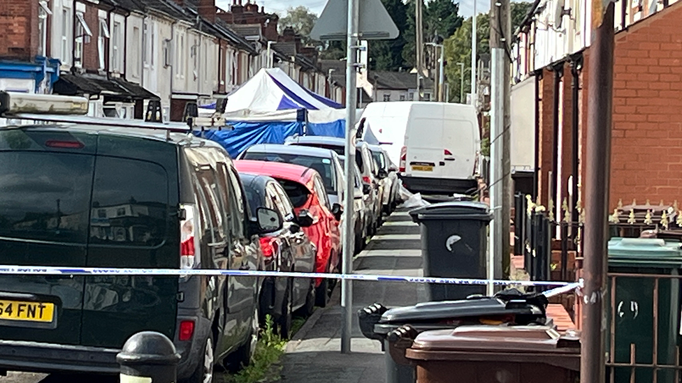 Terraced houses on both sides of the road, with cars on both sides of the road.  Blue and white police tape is in the foreground and a blue and white tent is in the middle of the road.