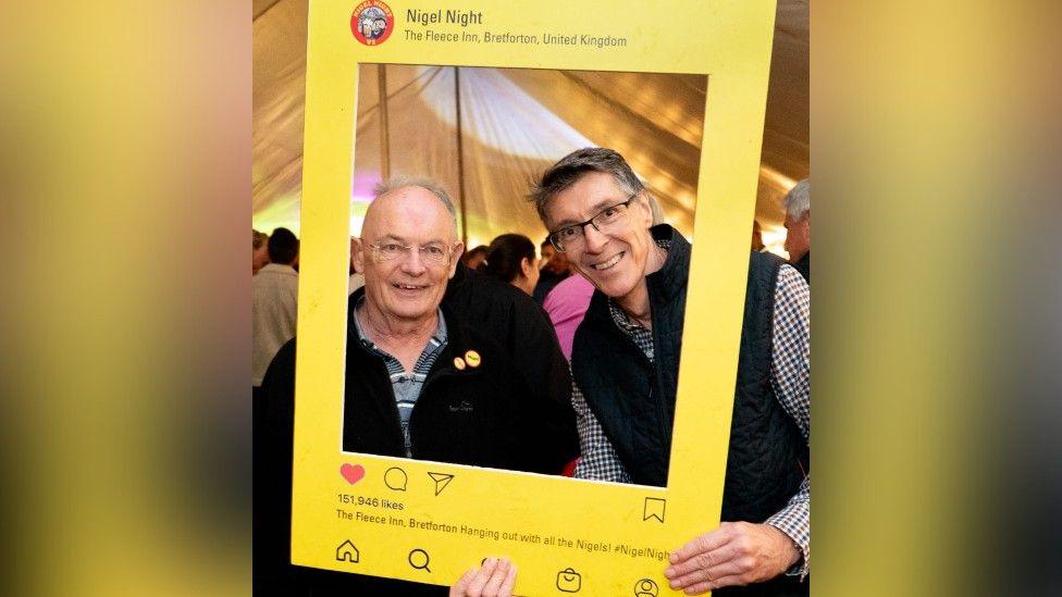 Two men pose for a photograph with their faces inside a large, yellow frame headed Nigel Night. They are in the marquee and there is a crowd behind them with lights reflected on the canvas.