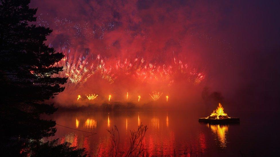 brightly coloured fireworks light up the sky as a firey beacon floats across a lake