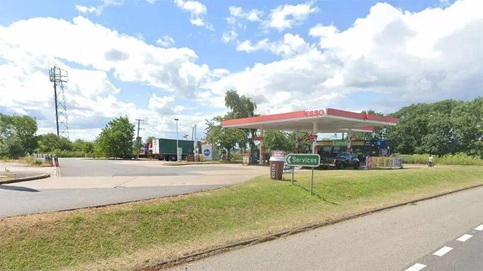 A petrol station forecourt at the side of a road, with a grassy embankment and lorries in the background. There is a green sign listing the available services.
