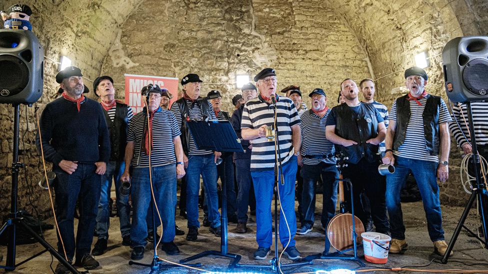 A band of men looking like sailors and holding tankards perform in a vault.