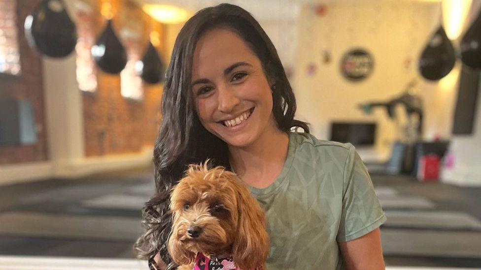 Leanne has long black hair and is wearing a green-t-shirt, she is smiling at the camera and holding a small dog.