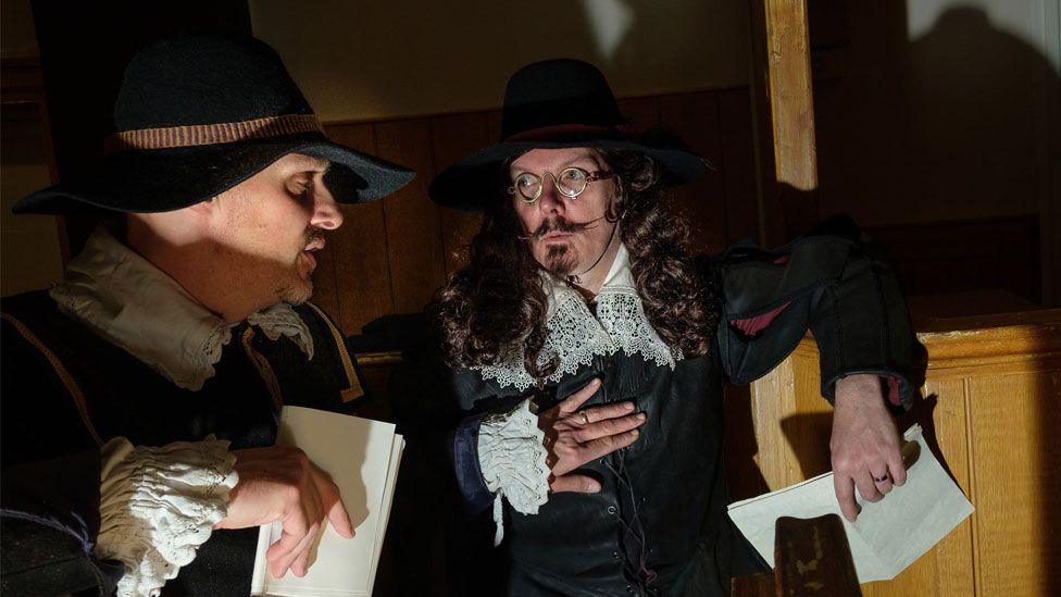 Two men in mid-17th Century dress during a re-enactment of the Huntingdon witch trial. They are wearing black broad-brimmed hats and dark clothing from which white linen collars and cuffs can be seen, decorated with broad lace. They are holding white documents and appear to be conferring.