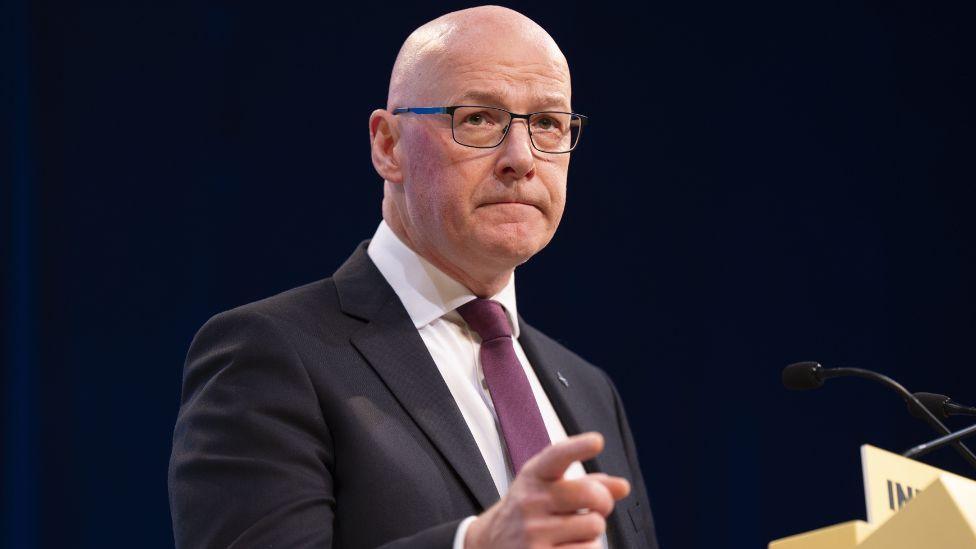 John Swinney, a bald man with glasses, points with his right hand while standing at a yellow podium. He is wearing a dark suit, white shirt and purple tie. 