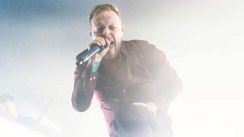 Sam Carter on stage. He's screaming into a microphone, with one leg elevated on a stage monitor in front of him. Bright white stage lights are flooding the space behind him.