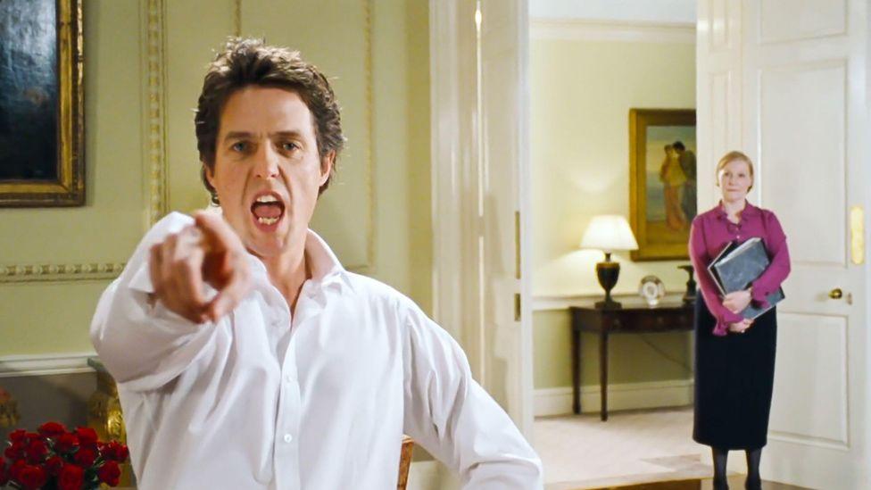 Hugh Grant, playing the prime minister, dances while pointing at the camera. He is wearing a white shirt with an open collar. Behind him a woman wearing a black skirt and a dark pink blouse, holding a file under her arm, watches him.