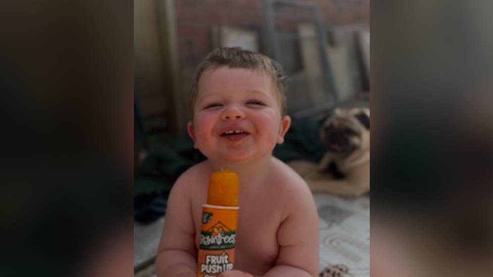 Picture of Osian smiling looking at the camera holding an orange ice lolly. He has short brown hair and is not wearing a top. He is sat in his garden. Behind him is a pug who is sat down.