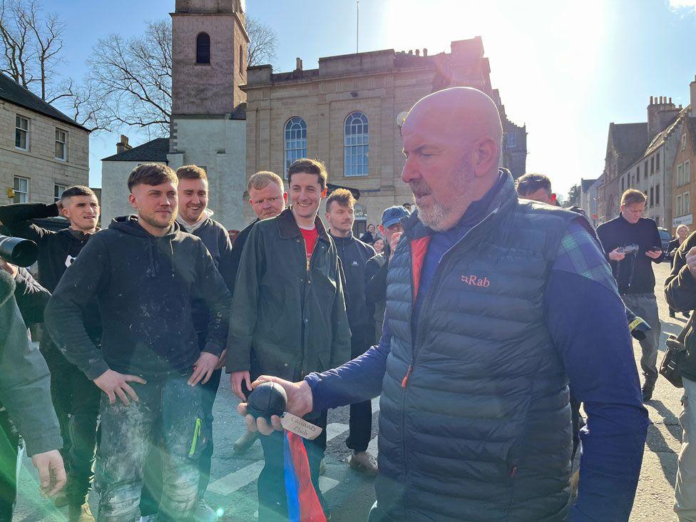 A man in a blue zip-up gilet holds a small leather ball with ribbons attached which he is about to throw into a group of men to start Jedburgh's ba' game