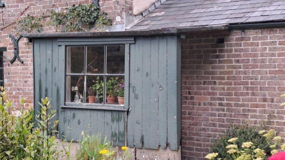 Wooden clad small porch attached to house with window in its centre - plants and flowers can been seen at its entrance.