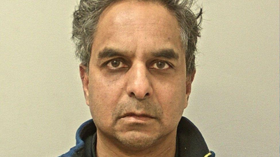 Police mugshot of Dr Amal Bose. He has short, unkempt dark hair that is greying and is looking directly at the camera, frowning slightly. He has brown eyes and bushy eyebrows.