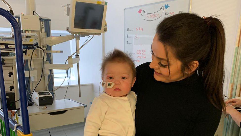 Baby Florence being held by mum Louise in her hospital room at GOSH. She is in a white sleepsuit and has a feeding tube. 