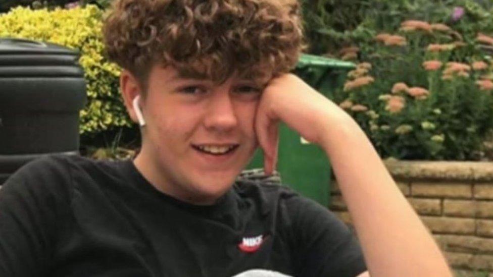 A smiling Olly Stephens with his head resting on his hand. He has curly brown hair and is wearing white earbuds and a black Nike t-shirt.