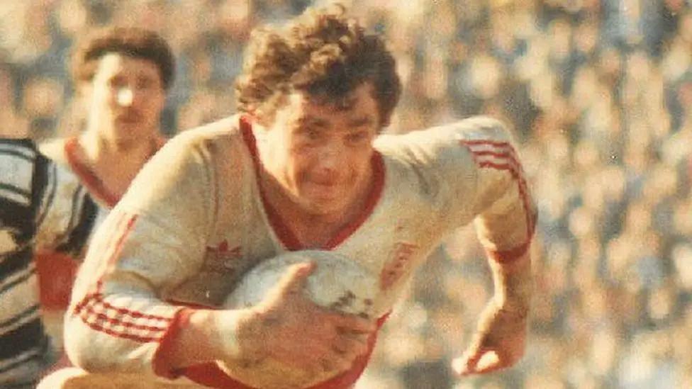 An archive image of Phil Lowe, wearing a red and white Adidas kit. He is carrying a rugby ball with a determined expression on his face. 