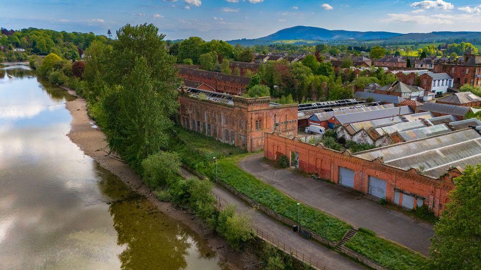 An aerial view of the disused Rosefield Mills building in Dumfries with other industrial buildings nearby and the reflection of trees in the River Nith