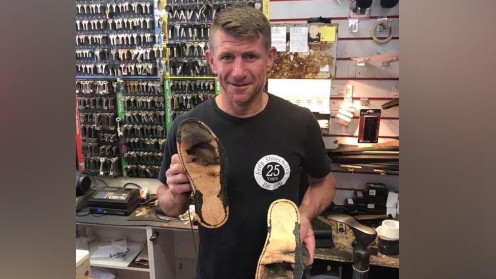 A man wearing a black T-shirt and holding a pair of damaged shoes in a workshop with racks of keys in the background.