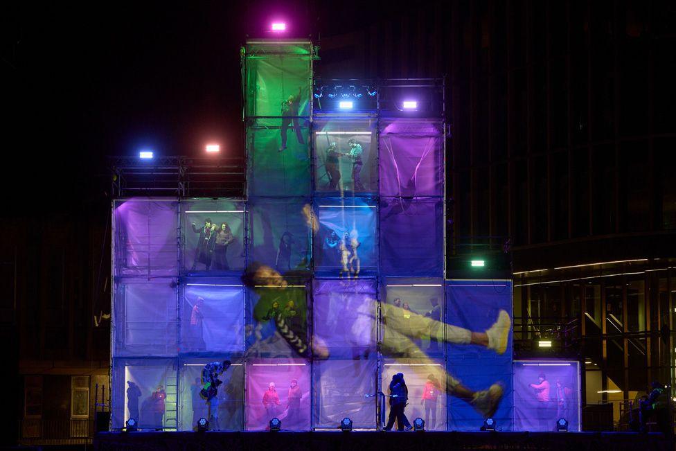 A shot of the stage showing towers made up of a series of colourful boxes with performers in them and a projection of a large figure across them all on the front