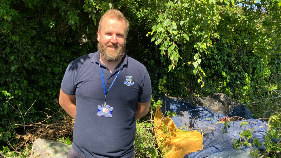 Sam Brunt in a polo shirt with bushes behind him