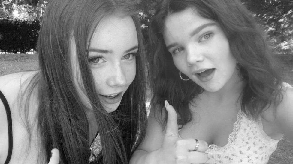 A black and white selfie of two teenage girls with long dark hair smiling and putting their thumbs up.