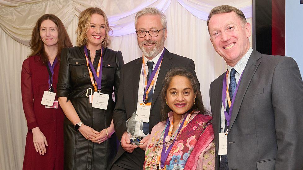 Lancashire County Council Trading Standards food team at the awards ceremony. Three women and two men, all dressed smartly, are wearing purple lanyards and smiling at the camera