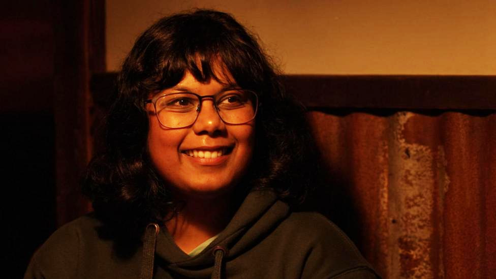 Swathi Padmanabhan smiling just at the right of the camera. She is wearing a dark green hoodie and is sitting in front of a brown and yellow wall. 