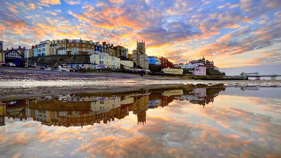 Cromer from the beach. The town rises up above the beach to the let in a curve, including a church tower and over to the right can be glimpsed its pier. Above the town is a blue sky, flecked with golden clouds from a sunset. The sky and town is crisply reflected in the sea